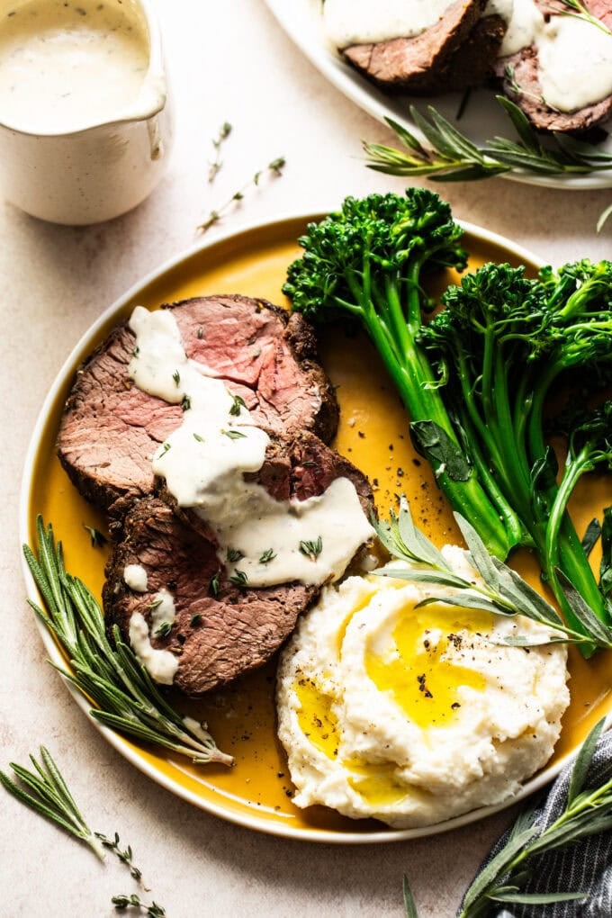 sliced beef tenderloin with horseradish cream sauce and mashed potatoes and broccoli on yellow plate